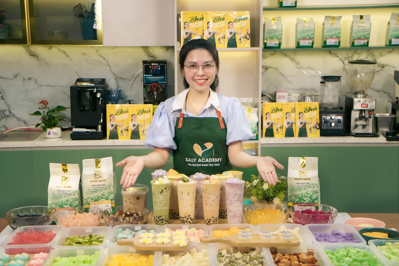 Giảng viên Ly Phạm đứng cạnh bàn nguyên liệu trà và bột sữa trong buổi quay khóa trà sữa